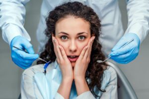Image of young woman sitting in dental chair with hands on her face in fear. Dental professional standing behind her with tools. root canal therapy dentist in Palmdale California
